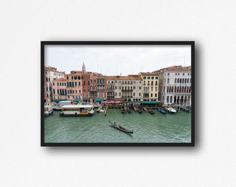 Puede incluir: Una vista de un canal en Venecia, Italia, con edificios coloridos que bordean la orilla del agua. Una góndola está en primer plano, con un gondolero remando por el canal. Hay varios otros barcos atracados a lo largo del canal.