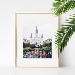 Puede incluir: Una fotografía enmarcada de un edificio blanco con altas torres, rodeado de árboles y una calle con carruajes tirados por caballos. El edificio está frente a un cielo nublado.