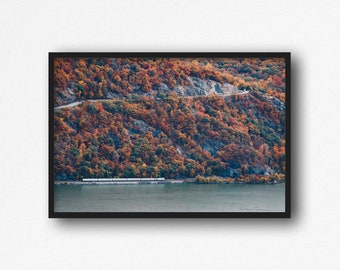May include: A train travels along a winding road through a forest with vibrant fall foliage. The trees are a mix of red, orange, yellow, and green. The train is white and has several cars. The river is calm and blue.