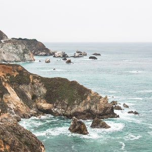 May include: A scenic view of a rocky coastline with a bridge in the distance. The ocean is a vibrant blue with white waves crashing against the rocks. The sky is overcast and gray.