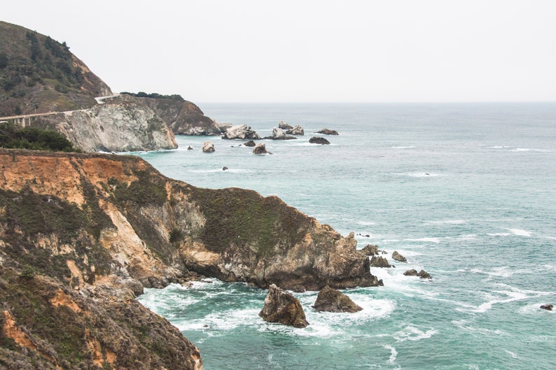 May include: A scenic view of a rocky coastline with a bridge in the distance. The ocean is a vibrant blue with white waves crashing against the rocks. The sky is overcast and gray.