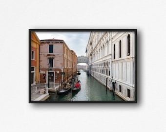 Puede incluir: Una vista de un canal estrecho en Venecia, Italia. El canal está bordeado de edificios a ambos lados, incluido un gran edificio blanco con ventanas arqueadas. Hay varias góndolas en el agua.
