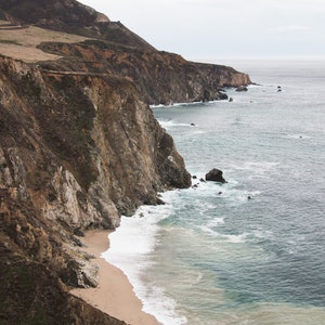 May include: A rocky cliffside with a sandy beach and foamy waves crashing on the shore. The ocean is a deep blue color and the sky is overcast.