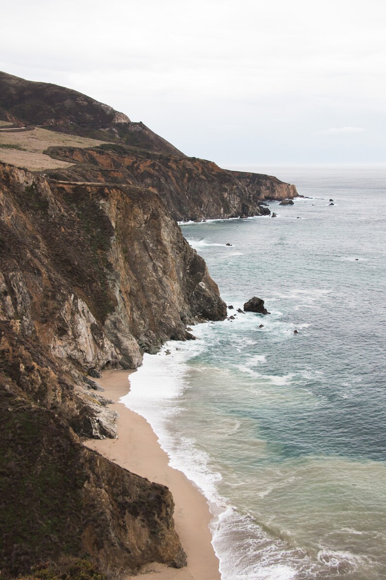 May include: A rocky cliffside with a sandy beach and foamy waves crashing on the shore. The ocean is a deep blue color and the sky is overcast.