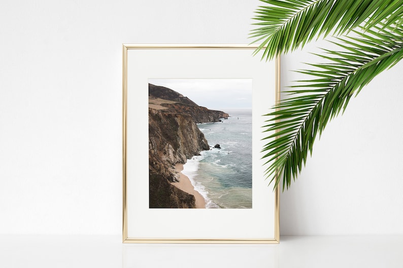 May include: A framed photograph of a rocky coastline with a sandy beach and blue ocean waves. The photo is in a gold frame and is hanging on a white wall.