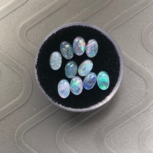 May include: A collection of oval-shaped opals in various shades of blue and white, displayed in a black velvet-lined container.