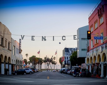 Peut inclure: Une scène de rue à Venice, en Californie, avec un panneau qui indique "VENICE" en grandes lettres. Le panneau est tendu à travers la rue entre deux bâtiments. Des palmiers bordent la rue et des voitures sont garées des deux côtés.