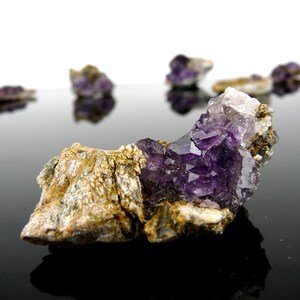 May include: A raw mineral specimen featuring deep purple amethyst crystals clustered on a textured, tan and grey rocky matrix. The cluster displays sharp, multifaceted crystal points with translucent edges, contrasting against the rough, earthy base.