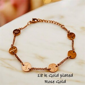 May include: Rose gold-plated bracelet featuring a chain design with six circular charms. Each charm has engraved Arabic script. The bracelet is described as 18k gold plated.