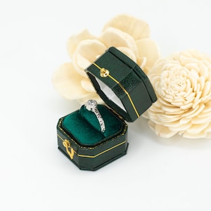 May include: A silver ring with a small round diamond in a green ring box with a gold trim.