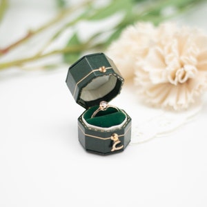 May include: An open, dark green, octagonal ring box with a gold clasp and trim, revealing a gold ring with a clear gemstone set on a green velvet interior. The box is on a white surface with a blurred floral background.