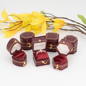 Peut inclure: Un ensemble de six petites boîtes à bagues bordeaux foncé de formes cœur, carrée et hexagonale, avec des bordures et fermoirs dorés. L'intérieur est doublé de tissu blanc et de velours rouge profond. Une boîte contient une bague argentée.