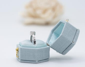 May include: A silver ring with a round gemstone sits in a light blue velvet ring box. The box is hexagonal and has a gold latch.
