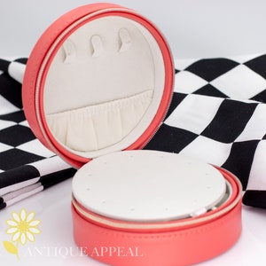 Pode incluir: Uma caixa de joias redonda de cor rosa coral com fecho de correr. A caixa est&aacute; aberta e o interior &eacute; forrado com tecido branco. A caixa tem um pequeno compartimento para brincos e um compartimento maior para an&eacute;is.