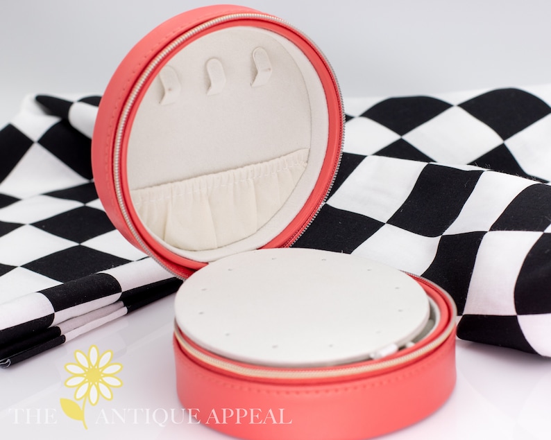 Pode incluir: Uma caixa de joias redonda de cor rosa coral com fecho de correr. A caixa est&aacute; aberta e o interior &eacute; forrado com tecido branco. A caixa tem um pequeno compartimento para brincos e um compartimento maior para an&eacute;is.