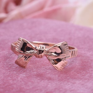 May include: A gold-toned ring featuring a bow design. The ring is displayed on a pink background.
