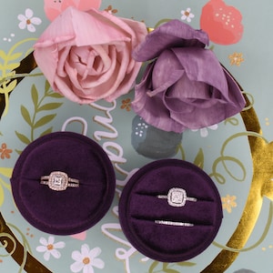Könnte beinhalten: Zwei purpurfarbene Samt-Ringboxen mit goldenen Bändern, jede mit einem Diamant-Verlobungsring. Die Boxen befinden sich auf einem hellgrünen Hintergrund mit goldenen und weißen Blumenmustern.