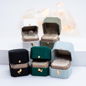 May include: An assortment of velvet ring boxes in various colors, including white, green, black, and blue. Each box is open, revealing a plush interior. One box contains a diamond ring. Gold-colored accents are visible.