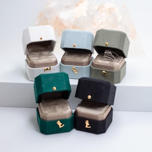 May include: A collection of hexagonal ring boxes in various colors: white, light blue, green, dark green, and black. Each box is open, revealing a velvet interior and a gold-colored clasp. Some boxes contain rings.