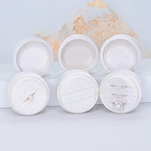 May include: Six white velvet ring boxes, round in shape, are displayed. Three boxes are open, revealing a ring in one and two rings in another. The boxes are arranged on a white surface, with a decorative backdrop.