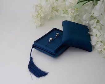 May include: A blue velvet ring box with a tassel, open to reveal two rings with diamonds. The box is sitting on a white surface with white flowers in the background.