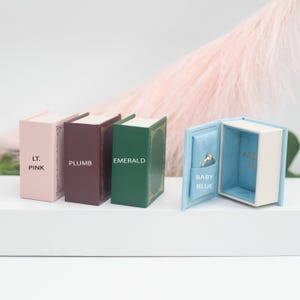 Puede incluir: Cajas para anillos con forma de libros en miniatura en rosa claro, ciruela, esmeralda y azul bebé. La caja azul bebé está abierta, revelando un anillo. Cada caja tiene texto en el lomo.