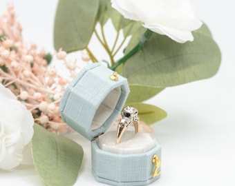 May include: A light blue ring box with a gold accent is open and a diamond ring is inside. The box is surrounded by white and pink flowers.
