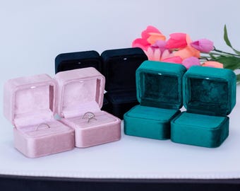 Könnte beinhalten: Geöffnete Ringboxen aus Samt in Rosa, Teal und Schwarz, einige mit Ringen. Im Hintergrund ein Strauß aus rosa und orangefarbenen Blumen. Die Boxen sind geöffnet und zeigen die Ringe.