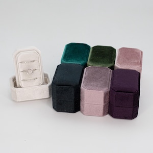 May include: A collection of seven velvet ring boxes in various colors including cream, teal, forest green, dusty rose, black, mauve, and deep purple. One cream box is open, displaying three silver rings with small clear stones on its insert.