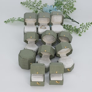 May include: A collection of sage green octagonal ring boxes with textured fabric exteriors and gold-toned metal latches. Each box features a light gray velvet interior lining designed to hold a ring, with one box containing a gold band.