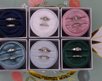 May include: Six velvet ring boxes in different colors, each containing two rings. The boxes are arranged in two rows of three. The boxes are white with a gold trim.