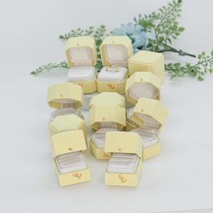 May include: A collection of pale yellow, octagonal ring boxes with textured fabric exteriors and cream-colored velvet interiors. Each box features a small gold-toned metal clasp on the front. One box contains a silver ring with a small stone.