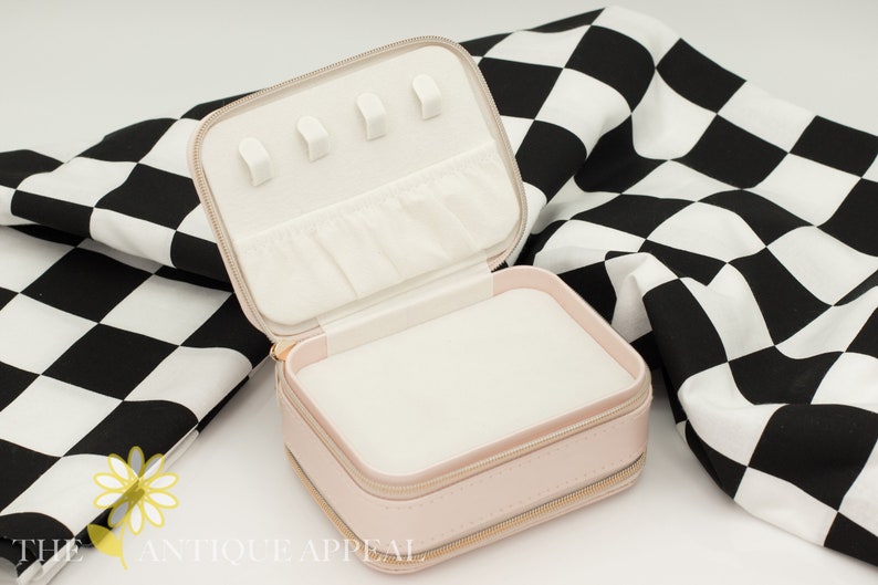 Pode incluir: Um estojo de viagem para joias rosa com fecho de correr. O estojo est&aacute; aberto e tem um interior branco com v&aacute;rios compartimentos para guardar joias. O estojo est&aacute; sobre um tecido xadrez preto e branco.
