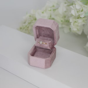 Pode incluir: Uma caixa de anel de veludo rosa pó com um anel de ouro dentro. O anel tem um único diamante redondo.