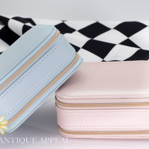 Pode incluir: Duas caixas de joias com z&iacute;per, uma azul claro e outra rosa claro, sobre uma superf&iacute;cie quadriculada em preto e branco. As caixas t&ecirc;m z&iacute;peres e costuras douradas.