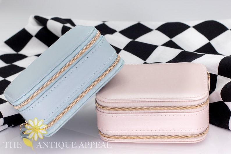 Pode incluir: Duas caixas de joias com z&iacute;per, uma azul claro e outra rosa claro, sobre uma superf&iacute;cie quadriculada em preto e branco. As caixas t&ecirc;m z&iacute;peres e costuras douradas.