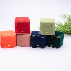 May include: An assortment of colorful, velvet-covered jewelry boxes in shades of red, orange, pink, blue, green, and light green. Each box has a gold-colored knob and a stitched design around the top. The boxes are arranged on a white surface.