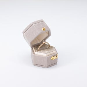 May include: A gold engagement ring with a small, clear gemstone sits inside an open, gray hexagonal ring box. The ring box has a gold clasp and a soft, beige interior. The ring has a delicate band with small, sparkling accents.