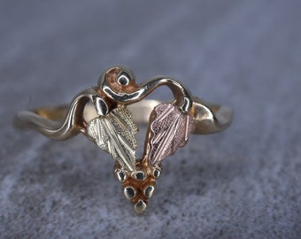 Puede incluir: Un anillo de oro con un diseño de tres hojas en diferentes colores de oro. Las hojas están conectadas por una vid y hay pequeñas cuentas doradas en la parte inferior de la vid.