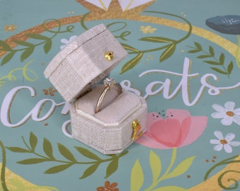 May include: A white ring box with a gold hinge and a gold inscription on the side. The box is open and a gold ring with a clear stone is inside. The box is sitting on a blue and green background with the word "Congrats" written in gold.