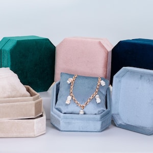 May include: A collection of velvet jewelry boxes in various colors, including teal, pink, and blue. One box holds a gold necklace with white charms. The boxes are octagonal and have a soft, plush appearance, ideal for storing jewelry.