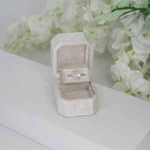 Puede incluir: Una caja de anillo de terciopelo blanco con un anillo de diamantes en su interior. La caja está sobre una superficie blanca con flores blancas en el fondo.