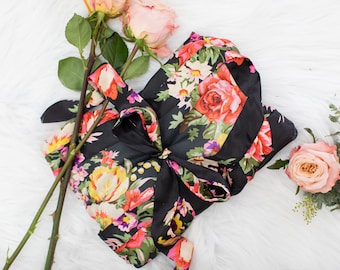 May include: A black silk robe with a floral print featuring pink, red, yellow, and green flowers. The robe is tied with a black sash.