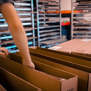 May include: A person's hands are seen arranging items inside open cardboard boxes. The boxes are brown and are in a row. Shelves filled with items are in the background. The scene suggests a warehouse or fulfillment center.