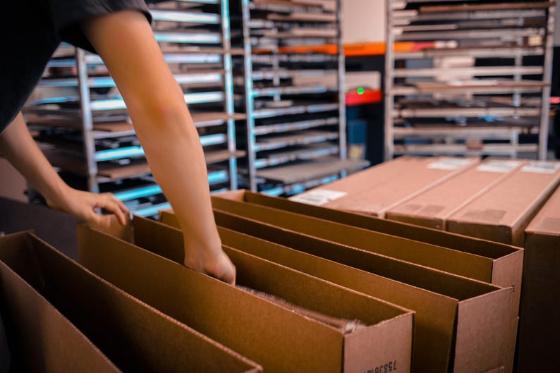 May include: A person's hands are seen arranging items inside open cardboard boxes. The boxes are brown and are in a row. Shelves filled with items are in the background. The scene suggests a warehouse or fulfillment center.