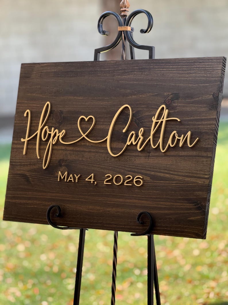 May include: A dark brown wooden sign with the names "Hope & Carlton" in gold script, with a heart symbol between the names. The date "MAY 4, 2026" is below the names. The sign is displayed on a black metal easel.