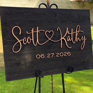 May include: A wooden wedding sign with the names "Scott & Kathy" in elegant cursive script, joined by a heart symbol. The date "06.27.2026" is displayed below. The sign is mounted on a black metal easel, set against a green lawn.