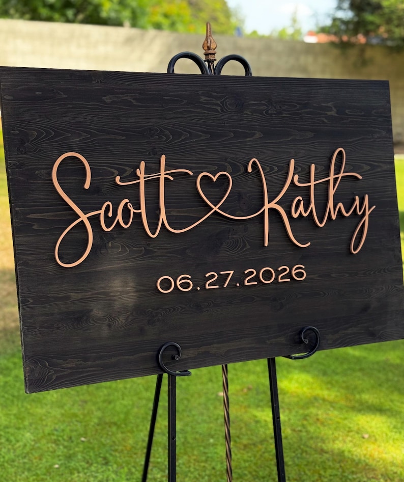 May include: A wooden wedding sign with the names "Scott & Kathy" in elegant cursive script, joined by a heart symbol. The date "06.27.2026" is displayed below. The sign is mounted on a black metal easel, set against a green lawn.