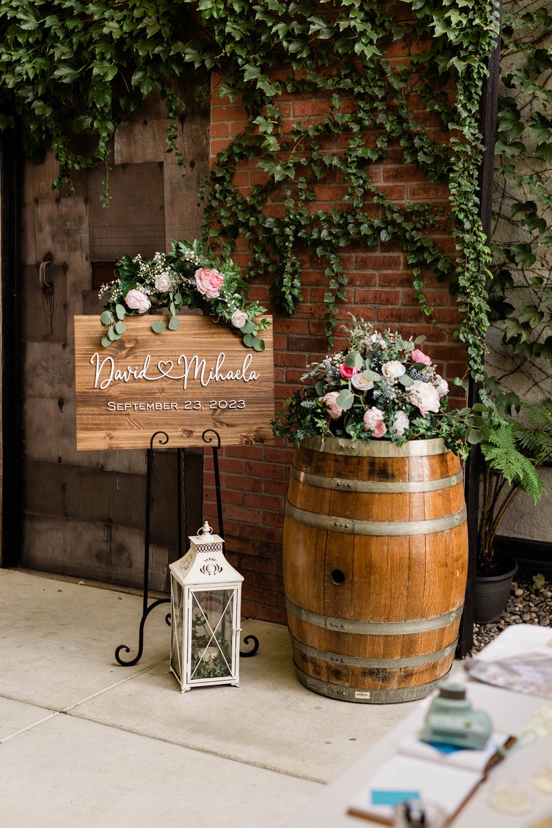 May include: A wooden wedding sign with the names "David & Mihaela" and the date "September 23, 2023" is displayed on a black metal stand. A white lantern and a wooden barrel with flowers are also present. Green vines and brick wall in the background.