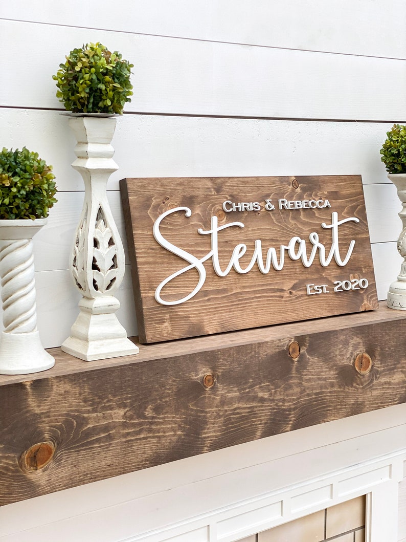 May include: A wooden sign with the text "Chris & Rebecca Stewart Est. 2020" in white raised letters. The sign is sitting on a wooden shelf with two white decorative candle holders on either side.